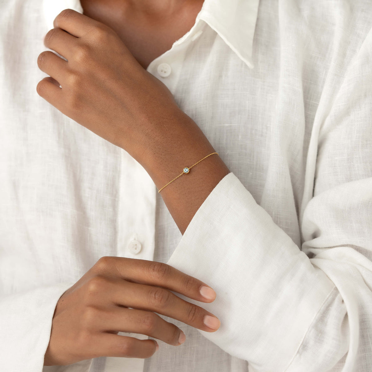 Close-up of a person wearing a white linen blouse, gently resting one hand while showcasing a delicate Gold Vermeil bracelet with a small Blue topaz gemstone centerpiece.