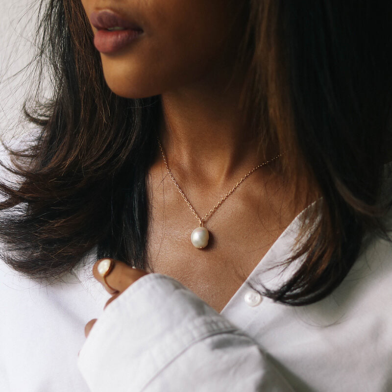 Close-up of a woman with medium brown hair wearing a gold chain necklace with a single pearl pendant and a pearl ring on her finger