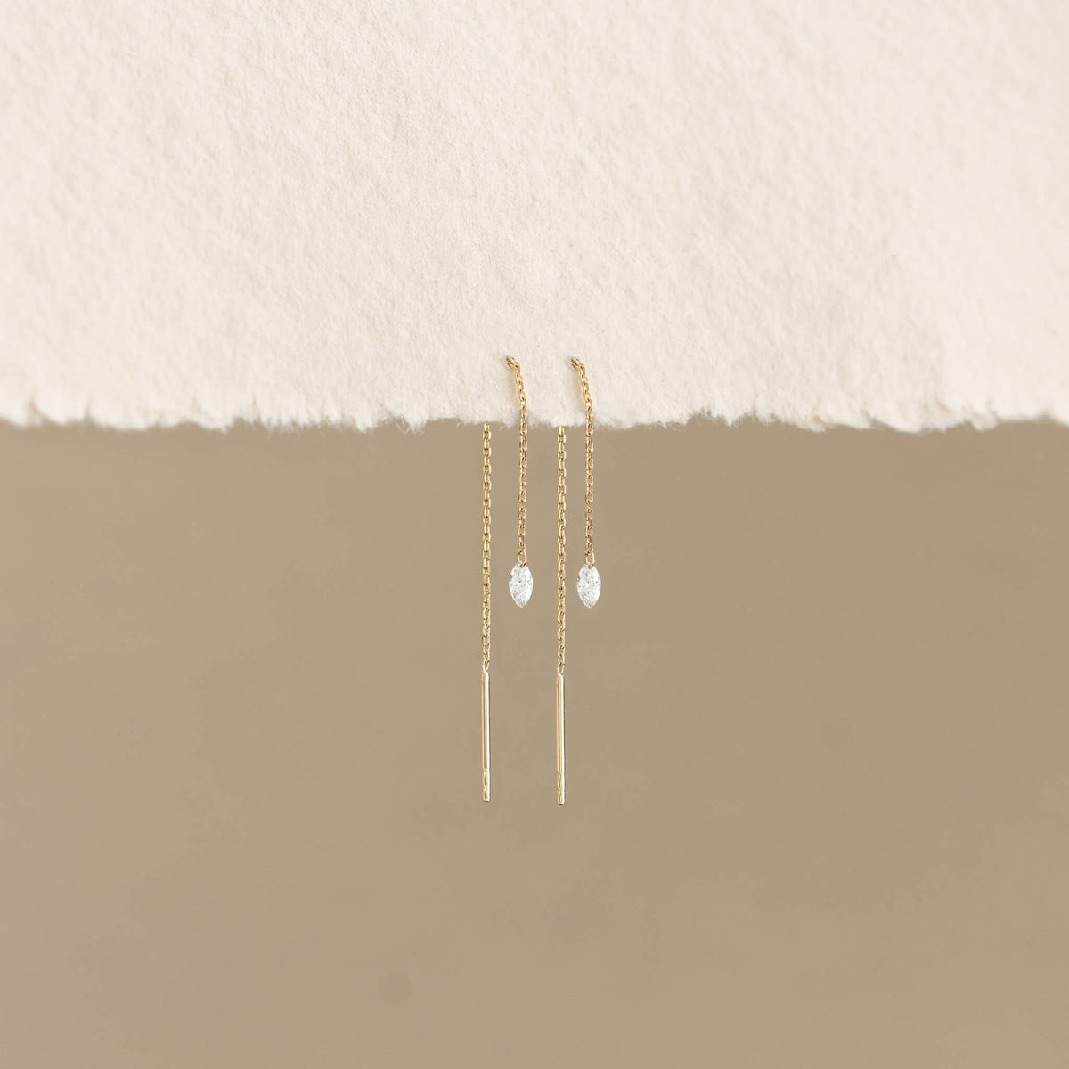 Elegant Gold chain earrings with teardrop gems hang against a beige background. Minimalist and delicate, they evoke a sense of simplicity and refinement.