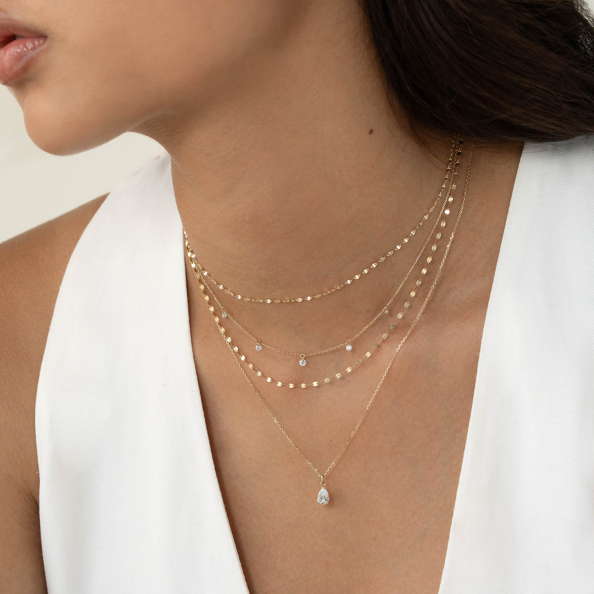 Close-up of a woman's neck adorned with layered gold necklaces featuring small, sparkling pendants. She wears a white top, conveying elegance and simplicity.