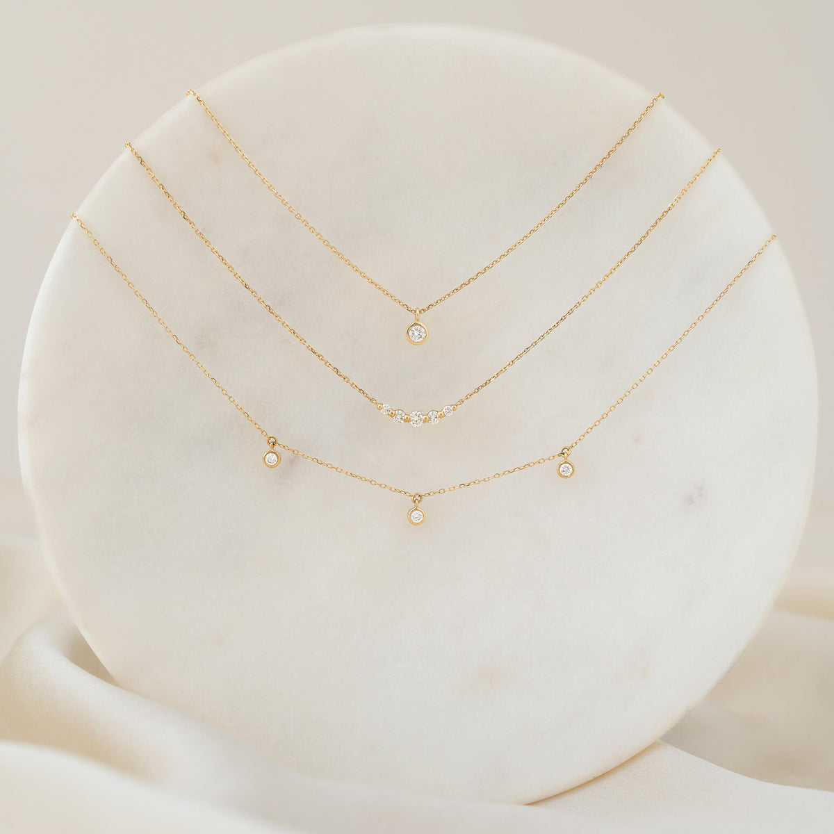 An elegant display of three delicate Gold necklaces layered on a round white marble stand. The top necklace features a single round diamond solitaire pendant in a bezel setting; the middle necklace is adorned with a gentle curve of five small diamonds; and the bottom necklace features three small bezel-set diamonds dangling at intervals along the chain.