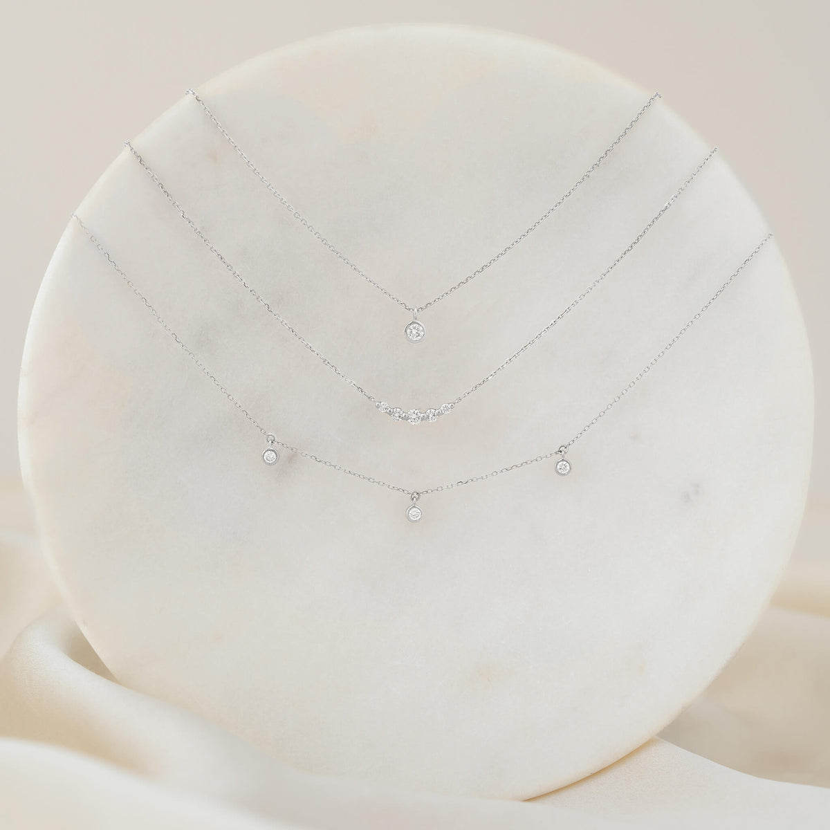 An elegant display of three delicate White Gold necklaces layered on a round white marble stand. The top necklace features a single round diamond solitaire pendant in a bezel setting; the middle necklace is adorned with a gentle curve of five small diamonds; and the bottom necklace features three small bezel-set diamonds dangling at intervals along the chain.