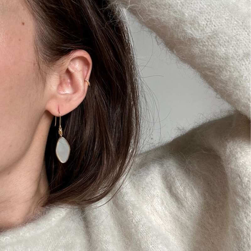 Close-up of a woman wearing a gold and white oval drop earring and a small gold hoop cuff against a neutral background