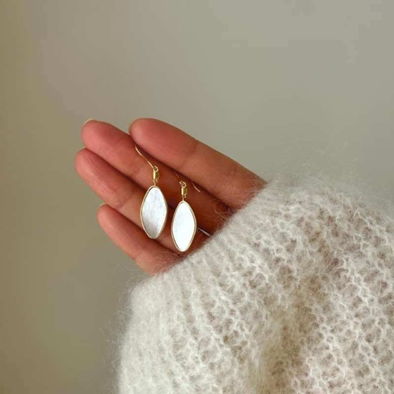 Close-up of a hand wearing a soft white knit sweater holding two gold hoop earrings with white marquise-shaped pendants