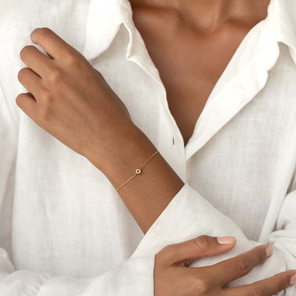 Close-up of a person wearing a white linen blouse, gently resting one hand on their shoulder while showcasing a delicate Gold Vermeil bracelet with a small Citrine gemstone centerpiece.