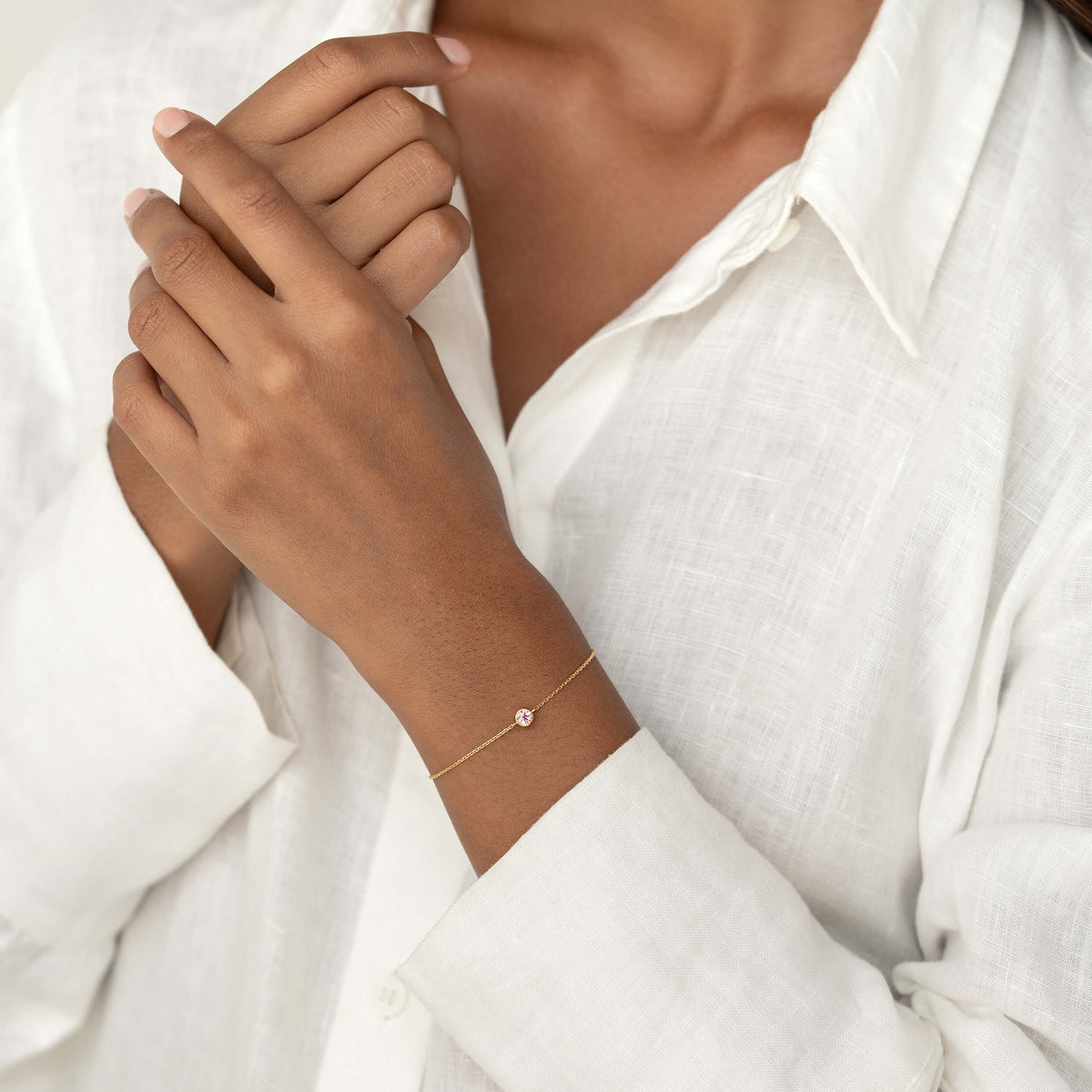 Close-up of a person wearing a white linen blouse, gently resting one hand on their shoulder while showcasing a delicate Gold Vermeil bracelet with a small Light pink tourmaline gemstone centerpiece.