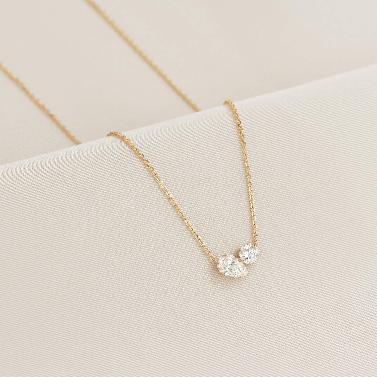 Delicate Gold chain necklace displayed on a soft, light fabric surface, featuring two diamond pendants placed side by side at the center.