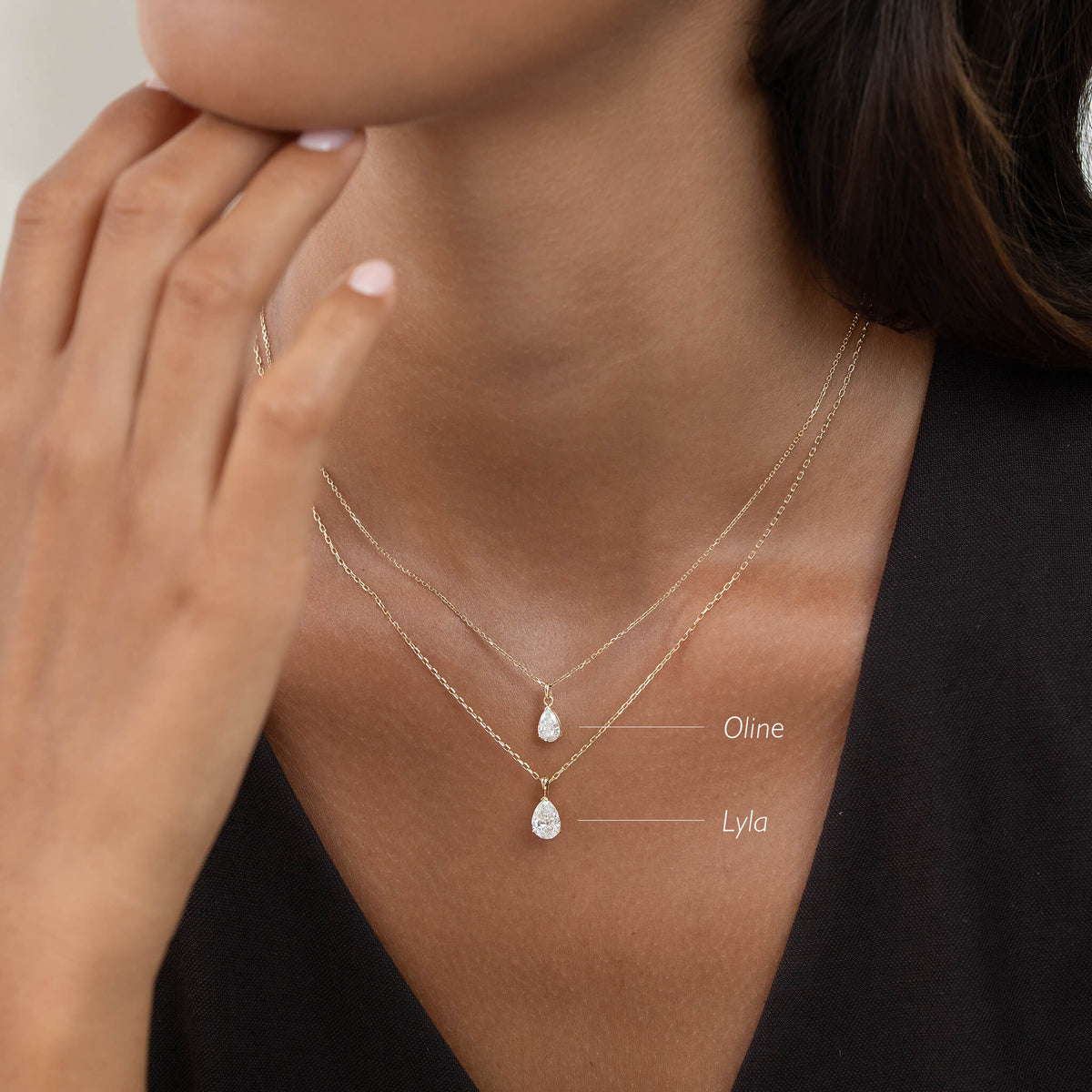 A close-up shot of a person wearing two delicate Gold layered necklaces, each featuring a pear-shaped diamond pendant. Text labels indicate the top, smaller pendant is named "Oline" and the lower, slightly larger pendant is named "Lyla."