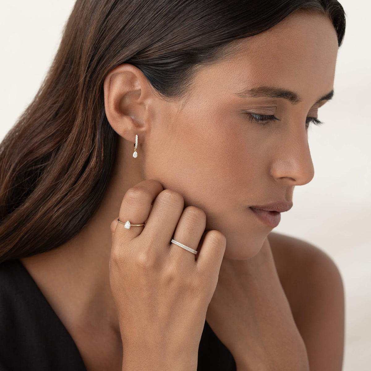 Woman wearing delicate Gold jewelry, including a band ring, pear-shaped gemstone ring, and matching drop earring, with her hand resting near her face against a soft neutral background.