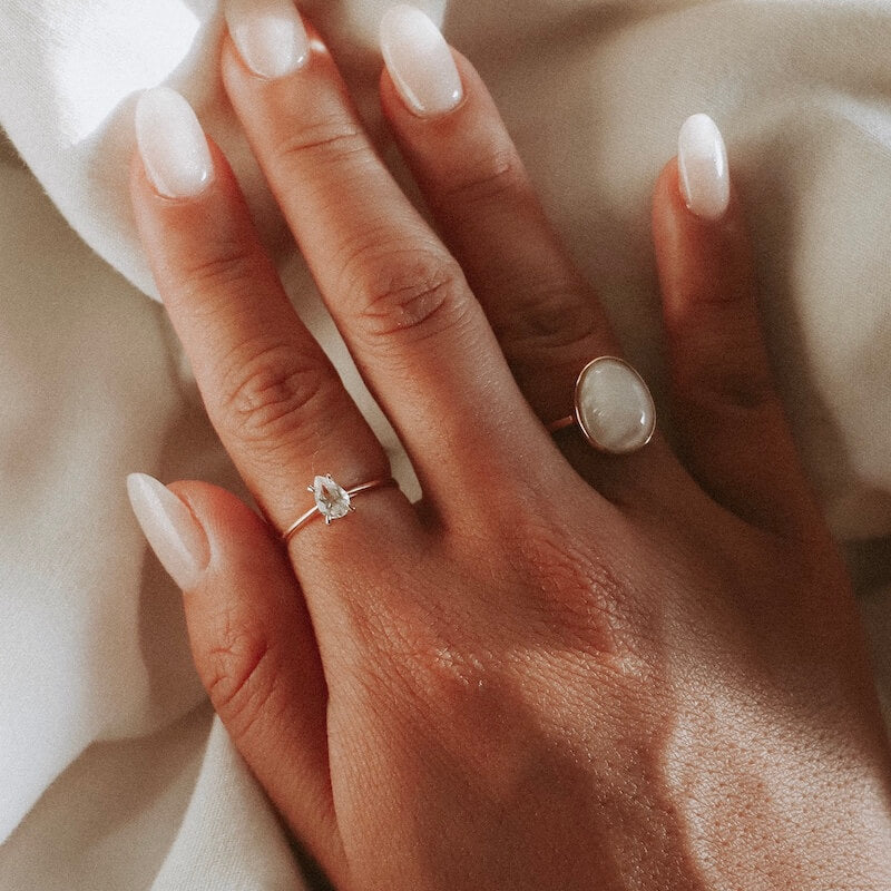 Close-up of a hand with oval and pear-shaped rings and natural manicured nails resting on soft beige fabric