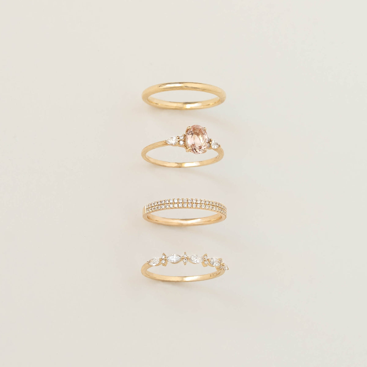 Four delicate Gold rings arranged vertically on a soft cream background, including a plain band, a pink gemstone ring with side stones, a double-row band, and a leaf-inspired diamond ring.