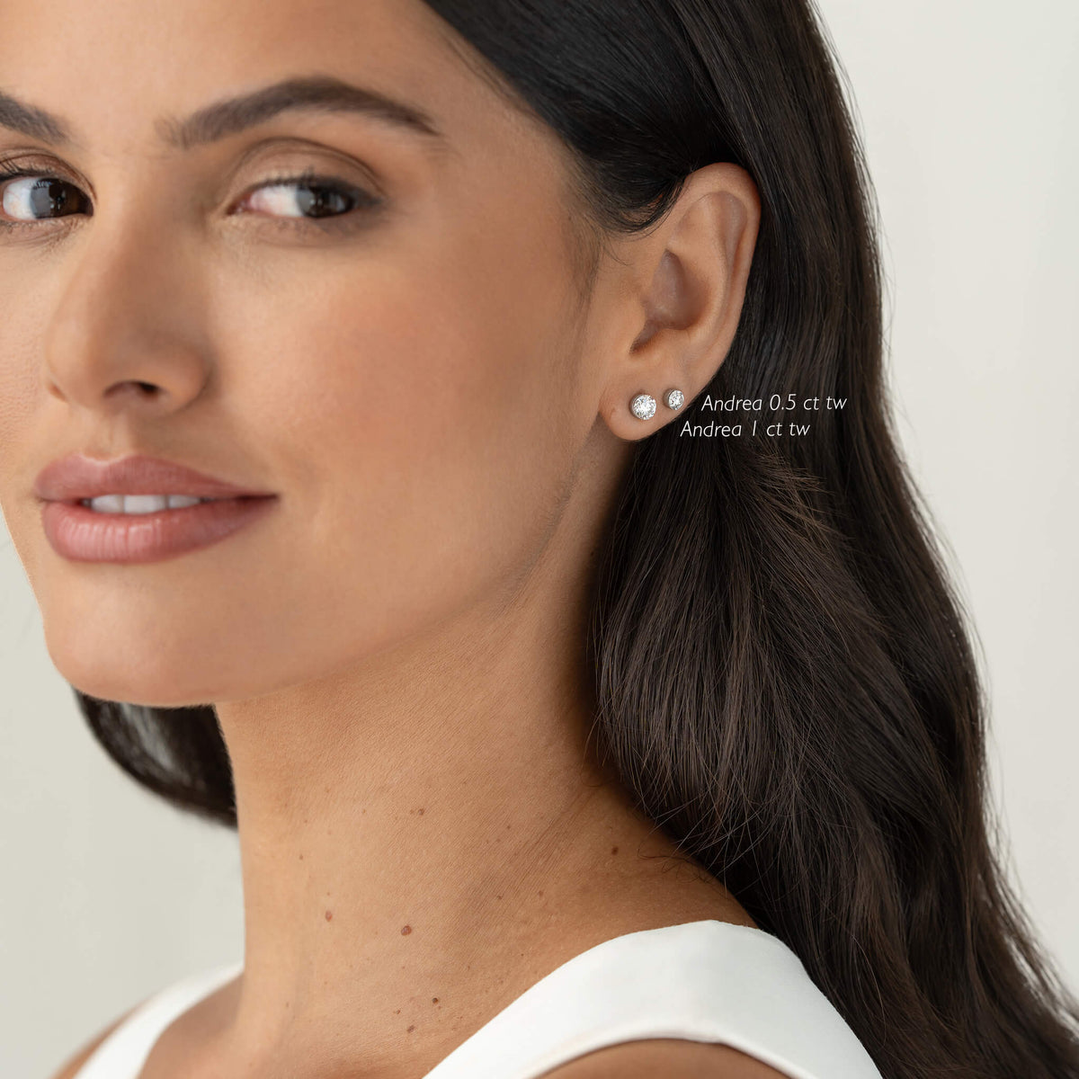 Close-up of a woman wearing two White Gold round diamond stud earrings on the earlobe, with small brilliant-cut stones labeled “Andrea 0.50 ct tw” and “Andrea 1 ct tw,” shown against a neutral background.