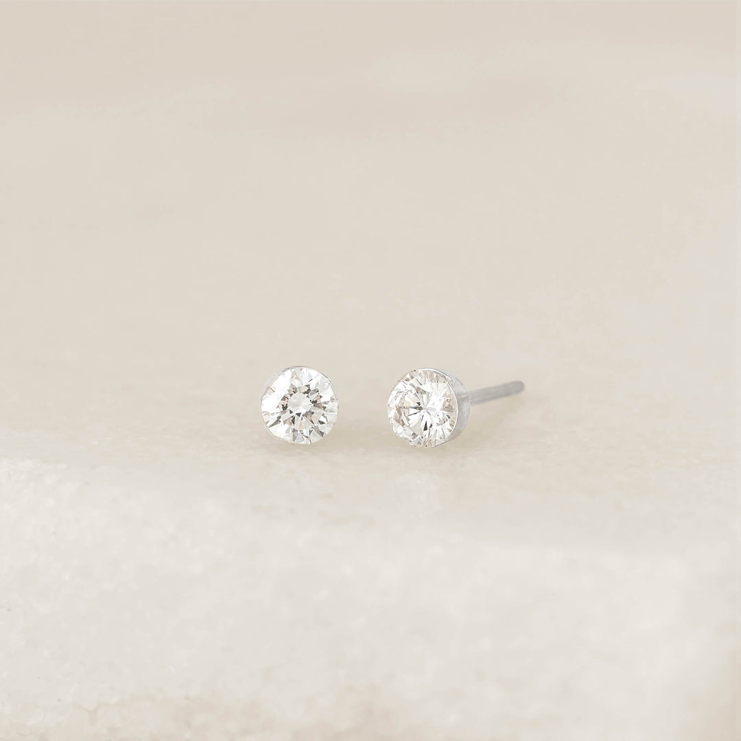 Pair of round White Gold diamond 0.50 ct tw stud earrings on a neutral surface, featuring brilliant-cut stones in simple settings with one earring showing the post.  | 0.5 ct tw