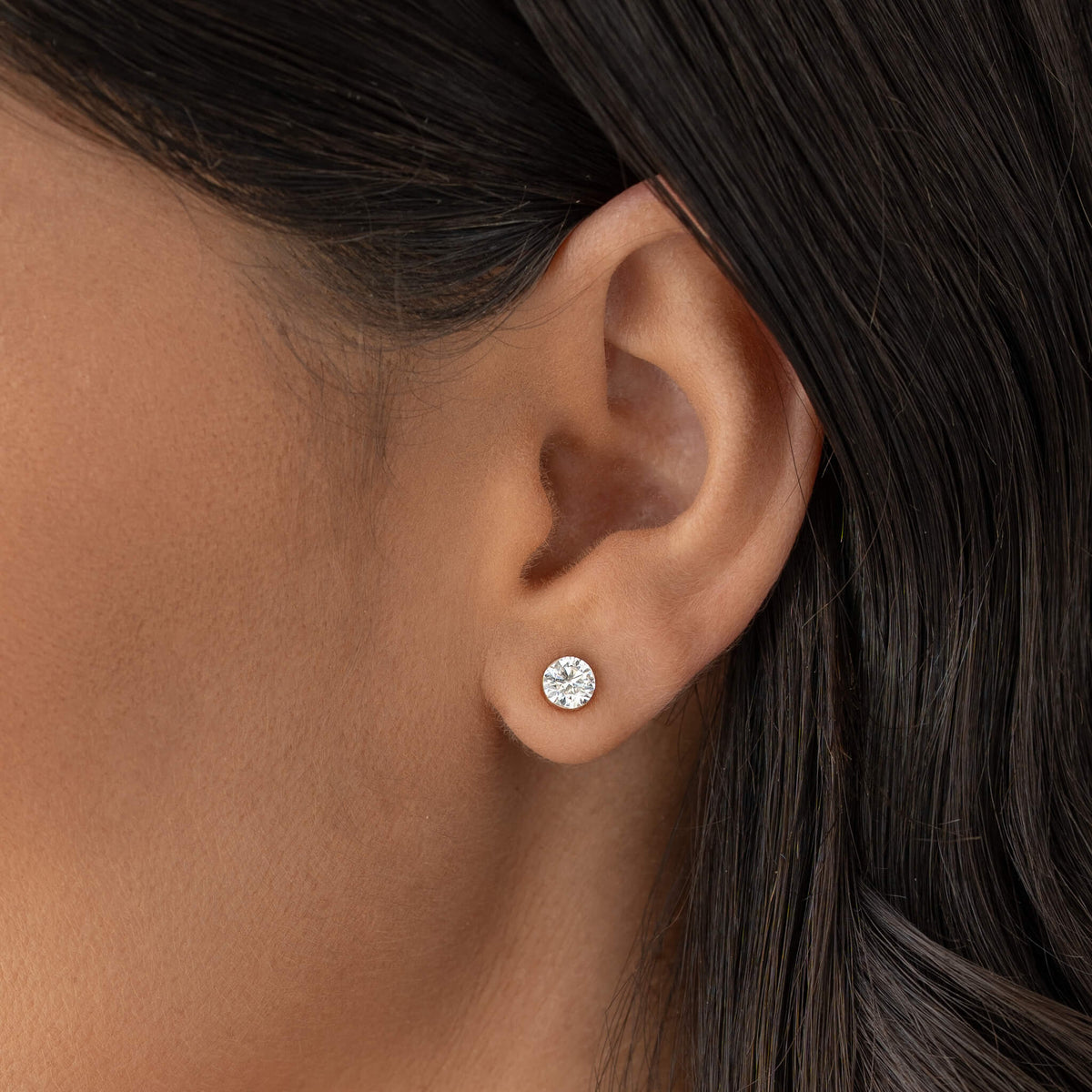 Close-up of a person’s ear with a small Gold 1 ct tw round diamond stud earring, set in a minimal setting, worn on the earlobe against smooth skin and dark hair background.