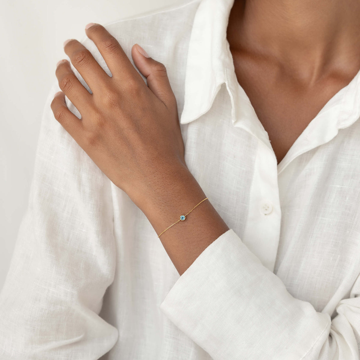 Close-up of a person wearing a white linen blouse, gently resting one hand on their shoulder while showcasing a delicate Gold Vermeil bracelet with a small London blue topaz gemstone centerpiece.