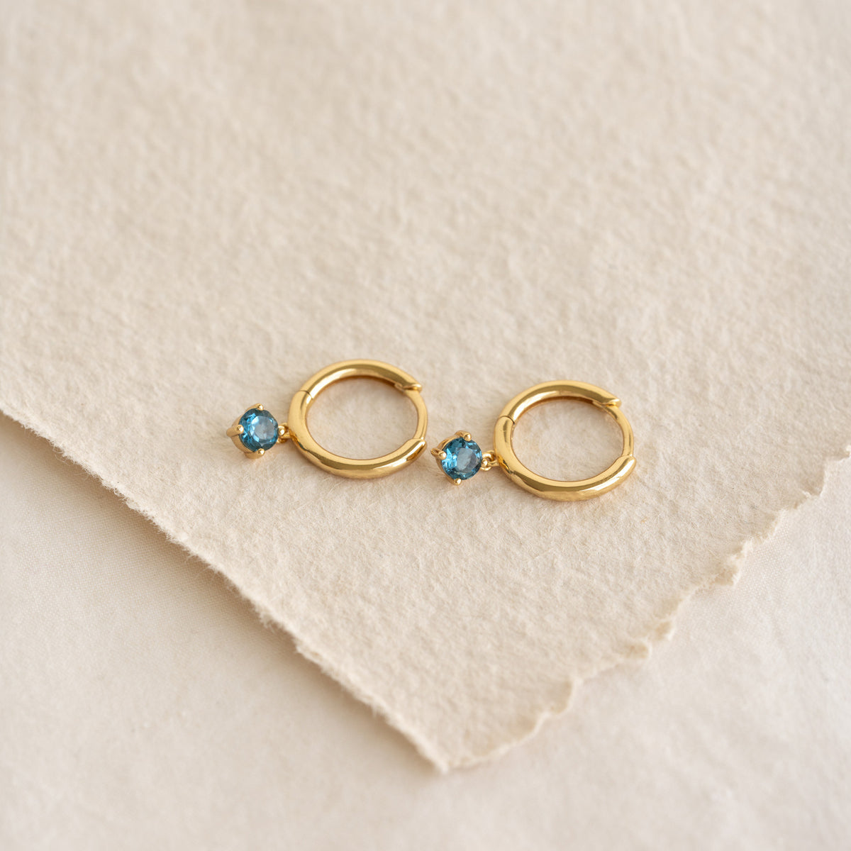 Close-up of September Birthstone Drop Huggie Earrings - London Blue Topaz with gold vermeil setting on textured paper background
