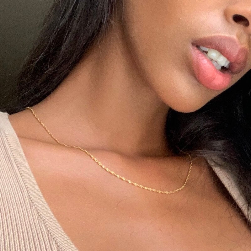 Close-up of a woman’s lower face and neck wearing a delicate gold chain necklace and a beige ribbed top against a neutral background