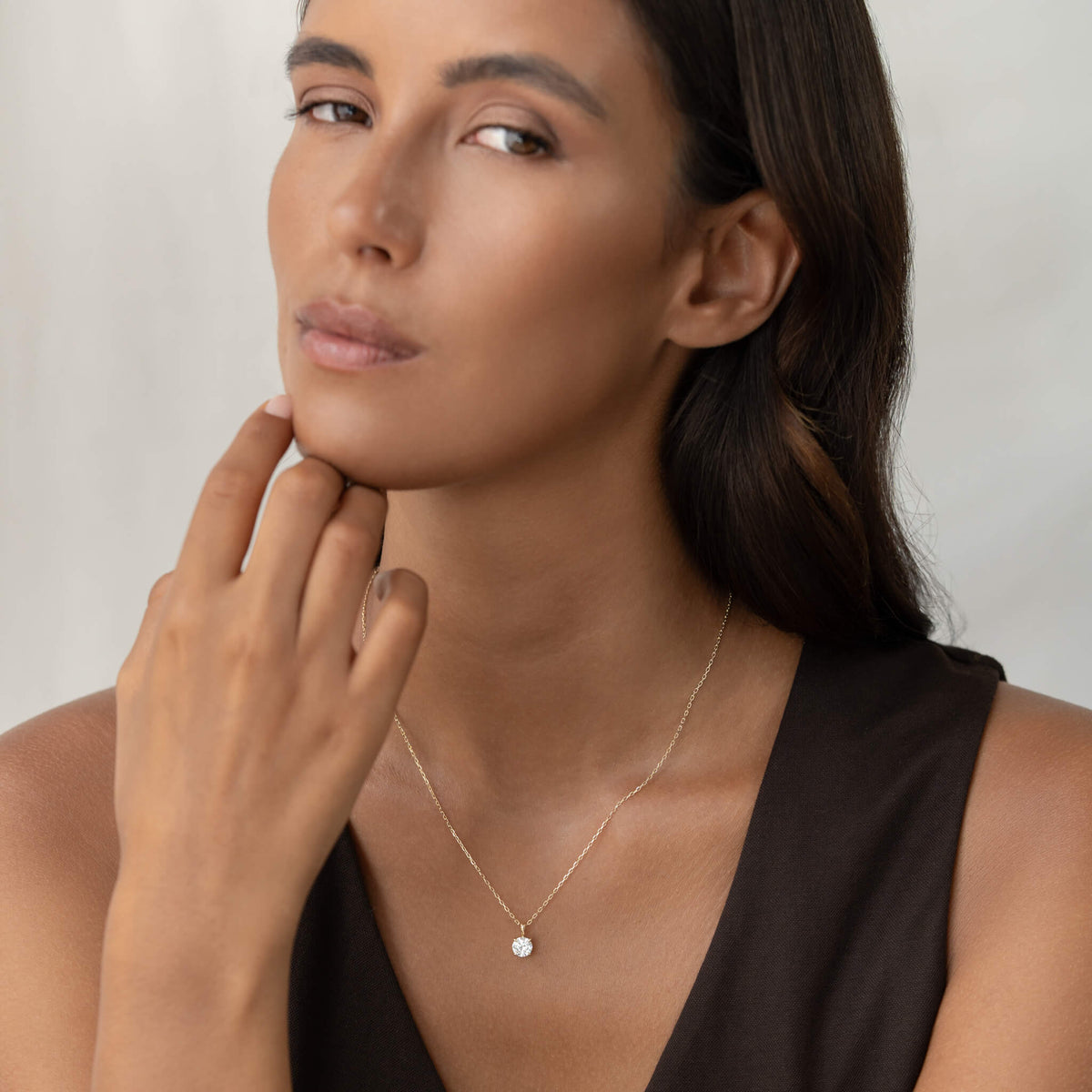 A portrait of a woman with dark hair wearing a delicate Gold chain necklace with a small round brilliant diamond solitaire pendant held in a prong setting, paired with a dark V-neck top.