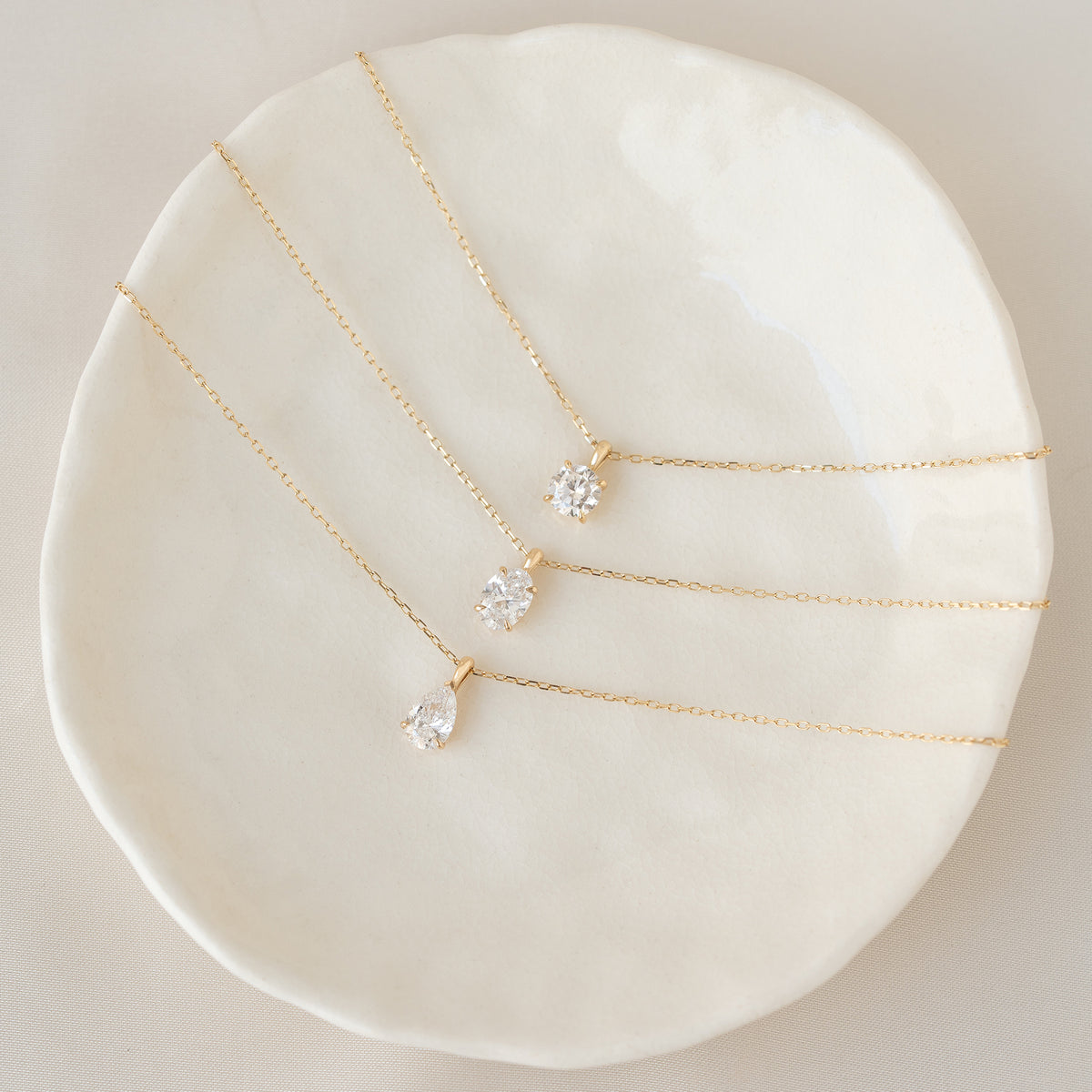 An overhead view of three delicate Gold necklaces arranged on a small, textured white ceramic dish. Each necklace features a different diamond cut: the top one has a round brilliant diamond, the middle one has an oval diamond, and the bottom one has a pear-shaped diamond.