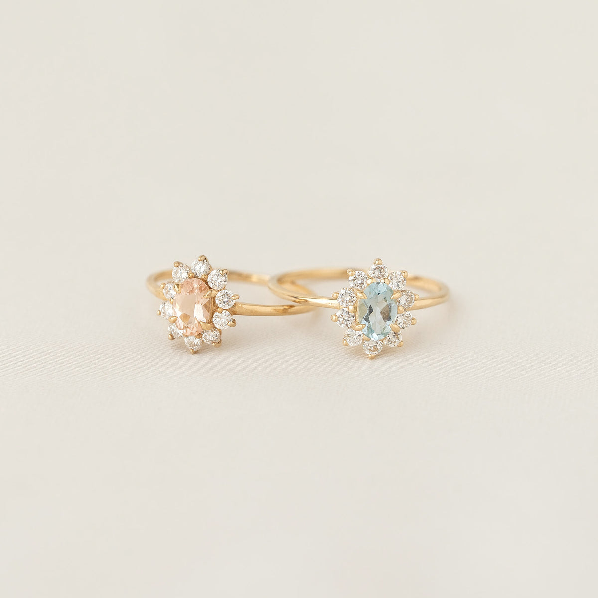 Two elegant gold rings with oval gemstones: one peach and one light blue, each surrounded by small sparkling diamonds, set against a soft beige background.