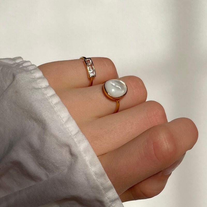 Close-up of a hand wearing two gold rings with white stone and rectangular gems against a neutral background