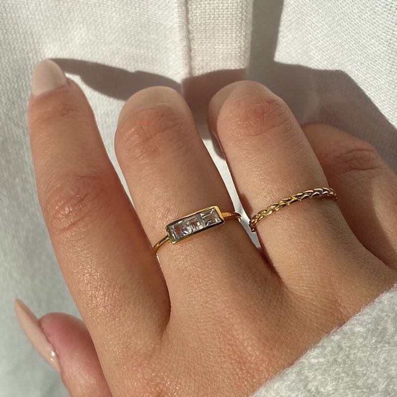 Close-up of a hand wearing two delicate gold rings against a soft white fabric background in natural light