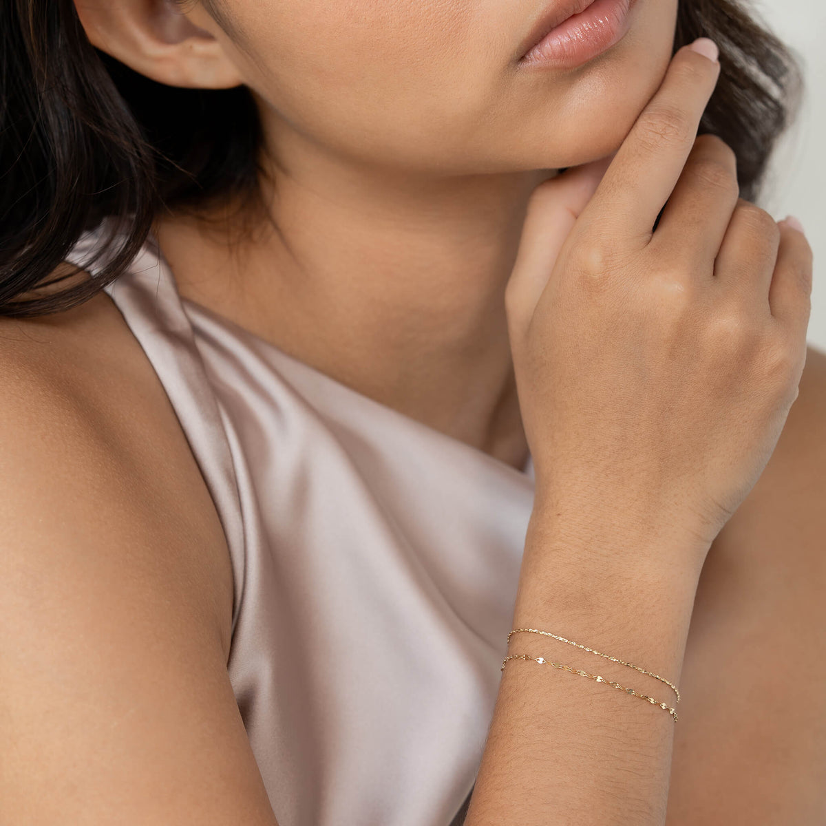 A woman wearing a silky sleeveless top gently touches her chin. She has delicate Gold chain bracelets on her wrist. The mood is elegant and contemplative.