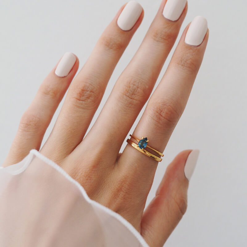 Close-up of a hand with pale pink nails wearing two gold rings, one featuring a small blue gemstone against a neutral background