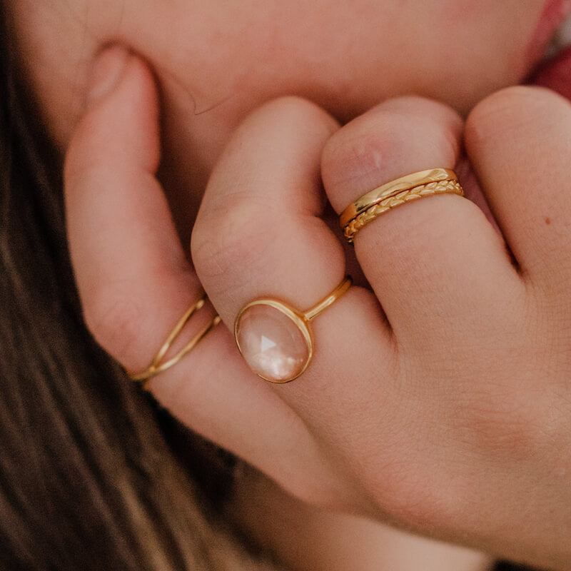 Close-up of a hand with three delicate gold rings, one featuring a polished oval stone, against a blurred background