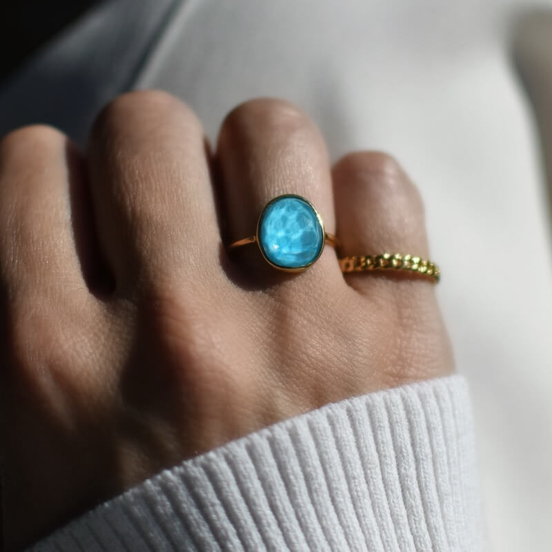 Close-up of a hand wearing a gold ring with a large blue stone and a textured gold band on a light fabric background