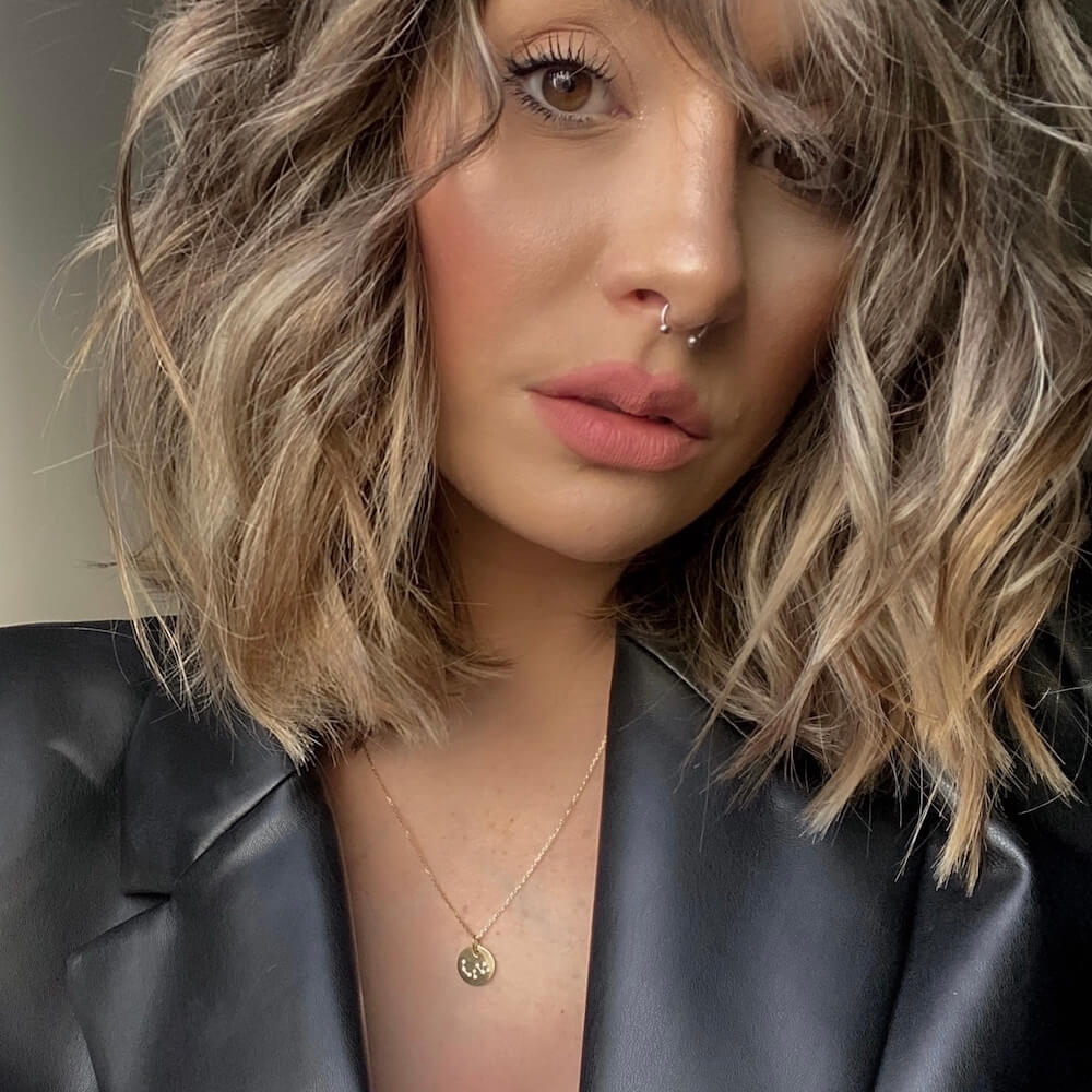 Close-up of a woman with wavy blonde hair wearing a black leather jacket and a delicate gold necklace with a pendant