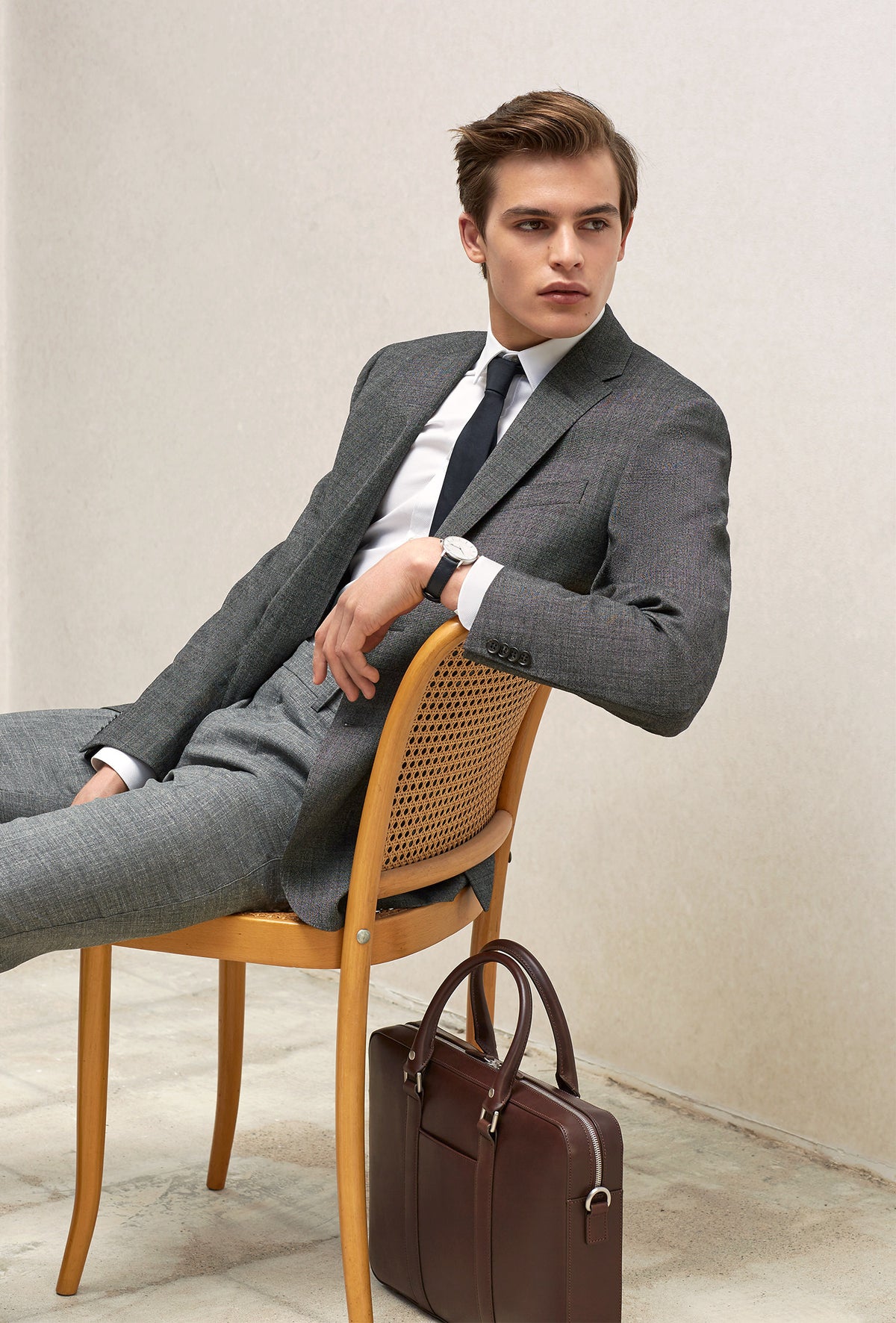 Man in grey suit sitting on wooden chair next to The Soft Briefcase (Private) in dark brown leather