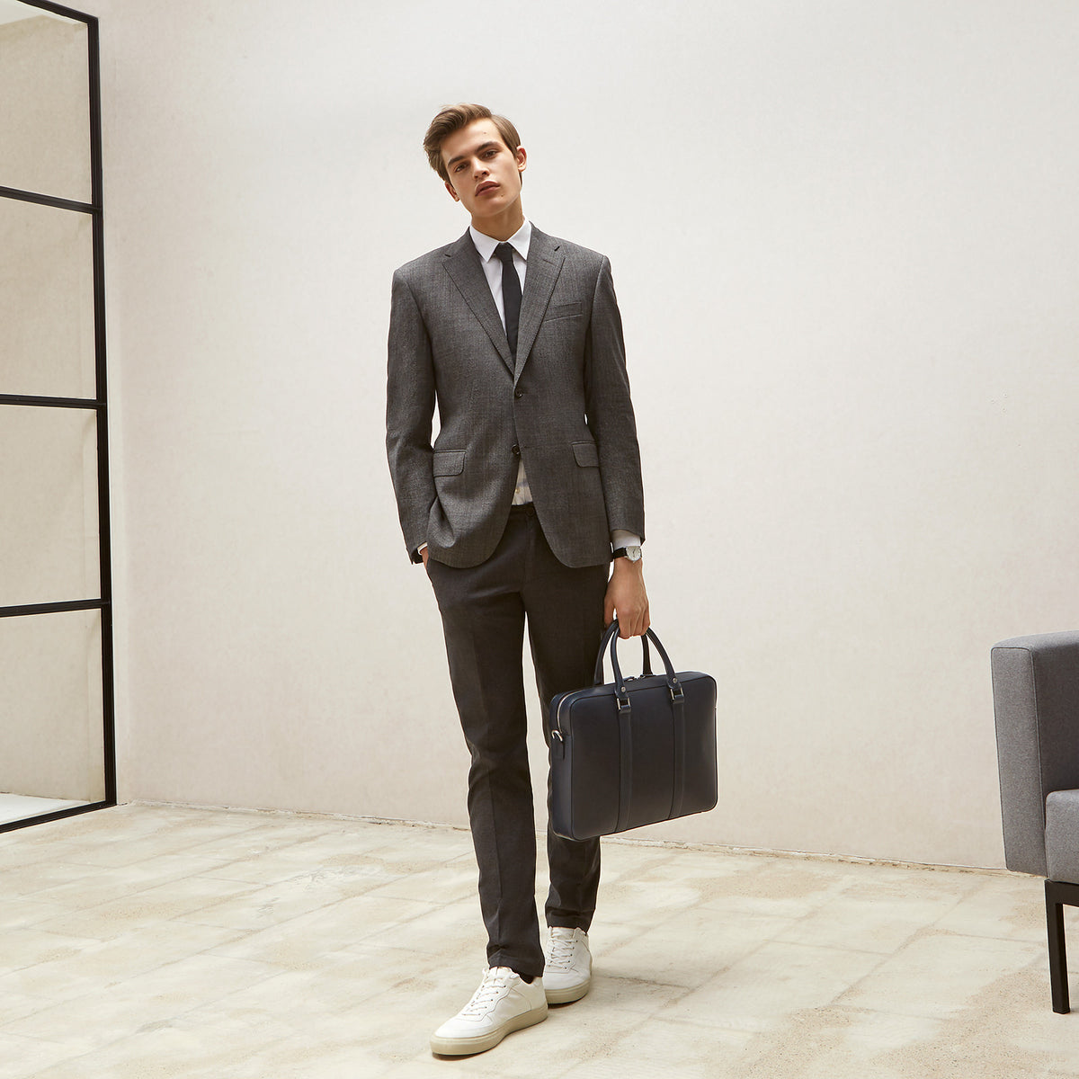 Man in tailored grey suit holding The Soft Briefcase (Private) leather briefcase standing in minimalist room