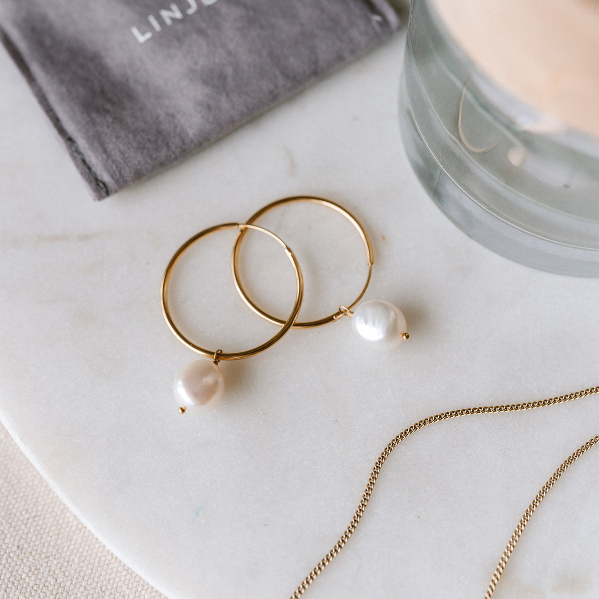Gold hoop earrings with white pearl drops resting on a light marble surface, styled beside a delicate gold chain and glass decor.